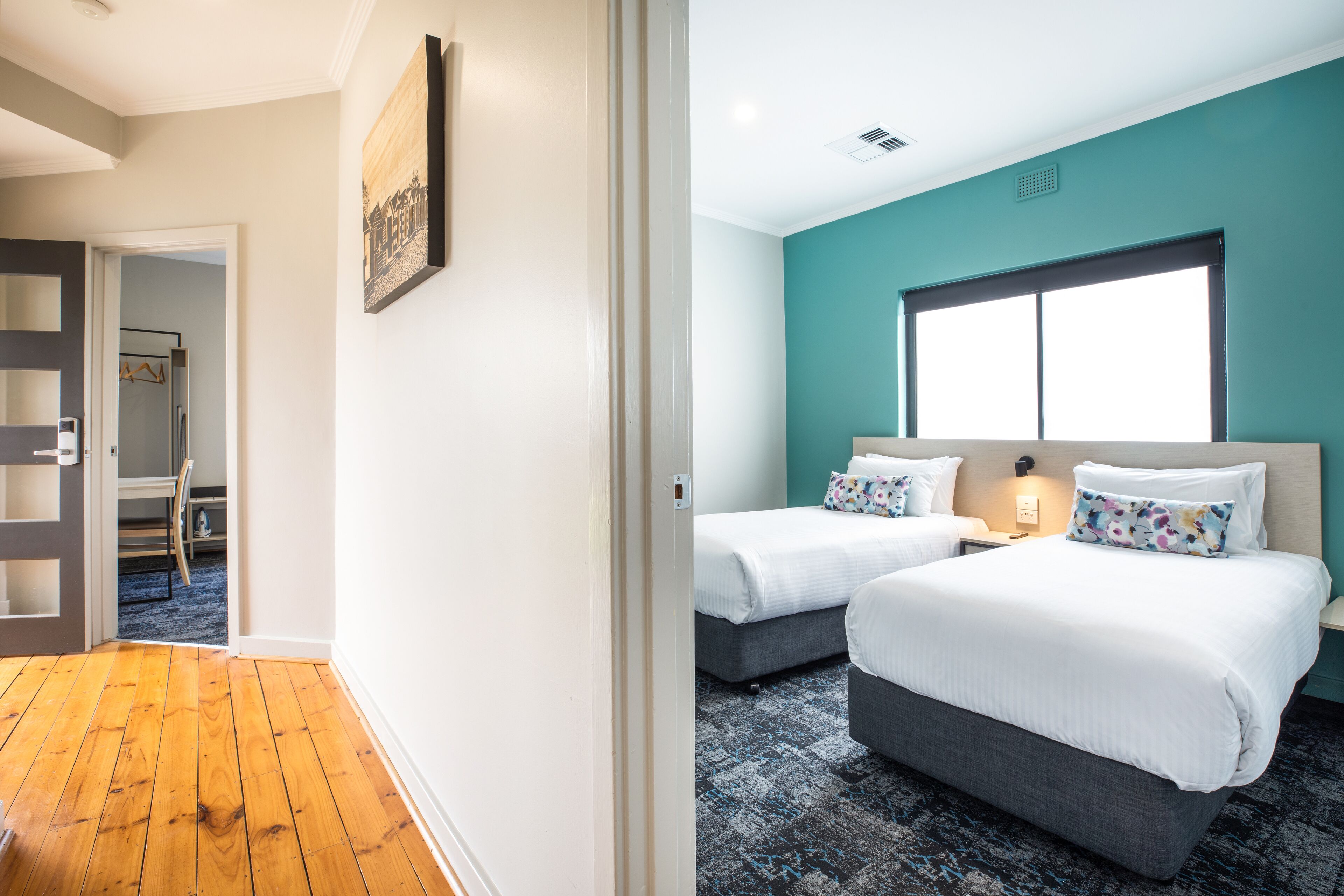 Quarto de hotel com duas camas, uma janela ampla e piso de madeira.