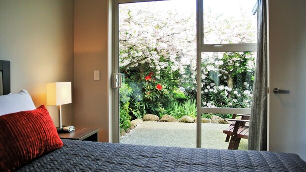 Een hotelkamer met een bed, een nachtkastje met een lamp en uitzicht op een tuin door een glazen deur.