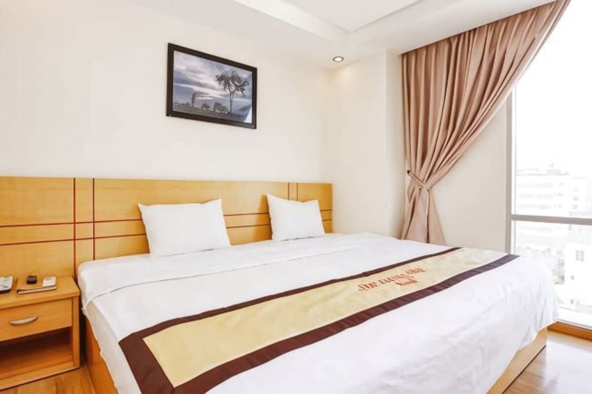 Une chambre d’hôtel avec un lit, une table de chevet, un tableau encadré au mur et une fenêtre avec des rideaux.