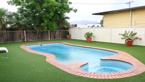 Una piscina rectangular con borde de ladrillo, rodeada de césped artificial y dos plantas en macetas.