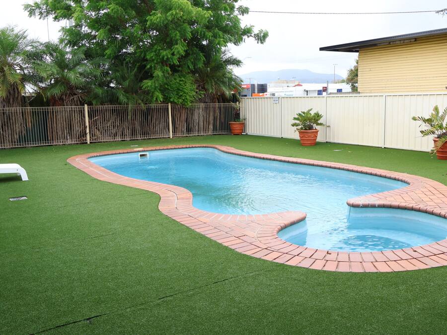 Uma piscina retangular com borda de tijolos, rodeada por grama sintética verde e duas plantas em vasos.