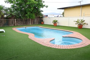 A rectangular swimming pool with a brick border, surrounded by artificial green grass, and two potted plants. - Sun Palms Motel (Allenstown)