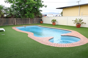 Una piscina rectangular con borde de ladrillo, rodeada de césped artificial y dos plantas en macetas.