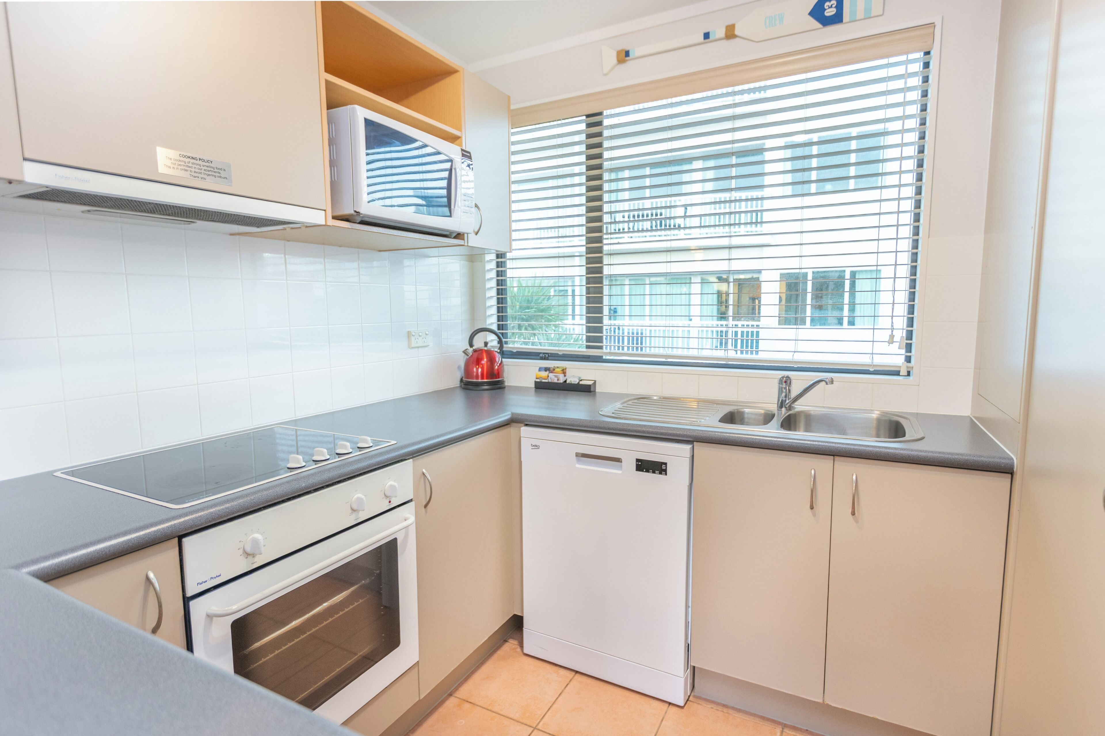 A modern kitchen with a built-in oven, a dishwasher, a microwave, and a window with blinds.