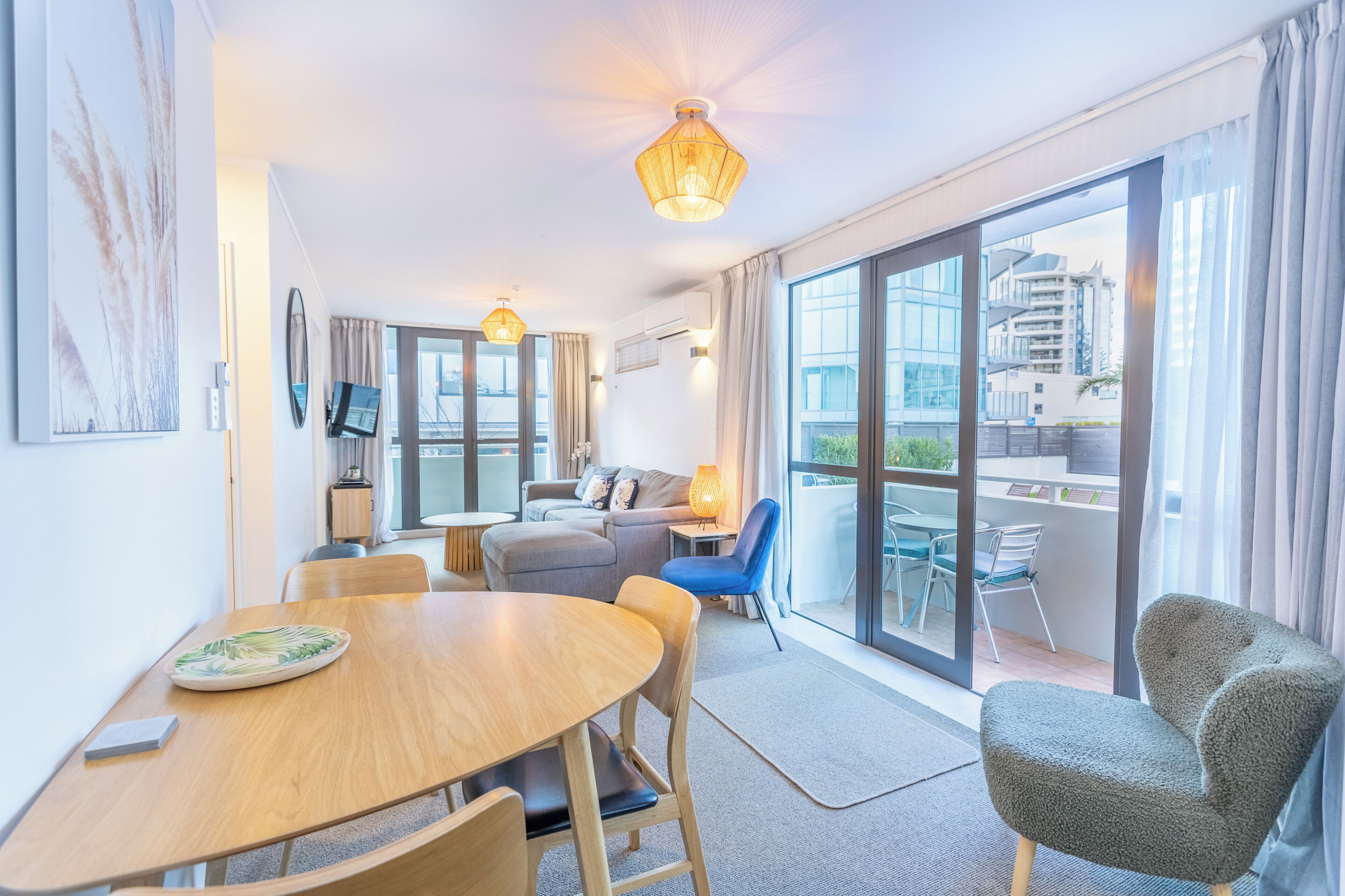 A modern hotel room with a dining area, a sofa, a balcony with a view of the city, and a large window.
