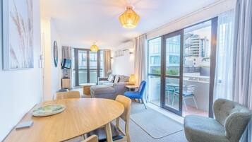 A modern hotel room with a dining area, a sofa, a balcony with a view of the city, and a large window.