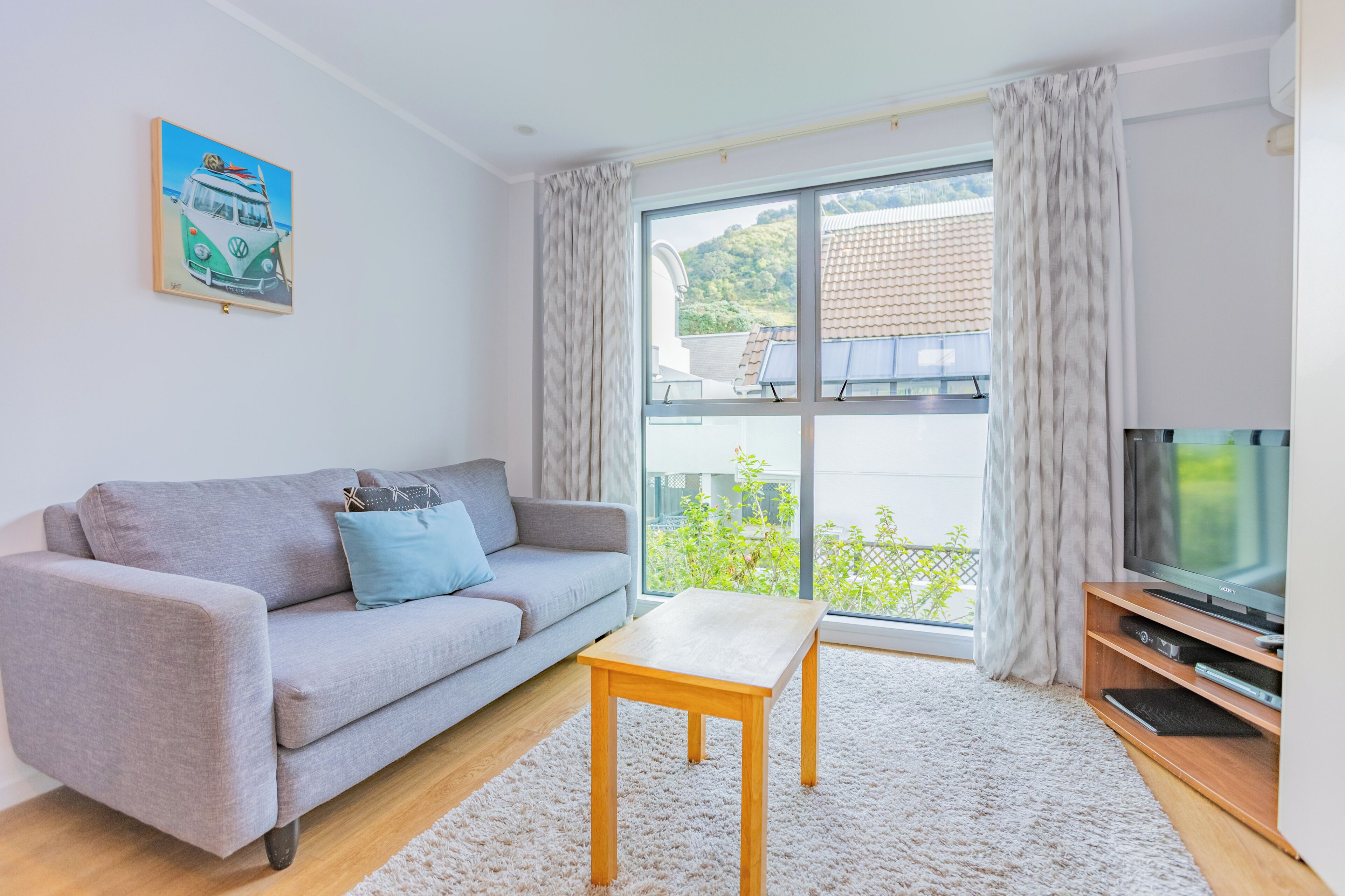 Uma sala de estar moderna com um sofá cinzento, uma mesa de centro de madeira, uma televisão de tela plana e uma janela grande com cortinas.