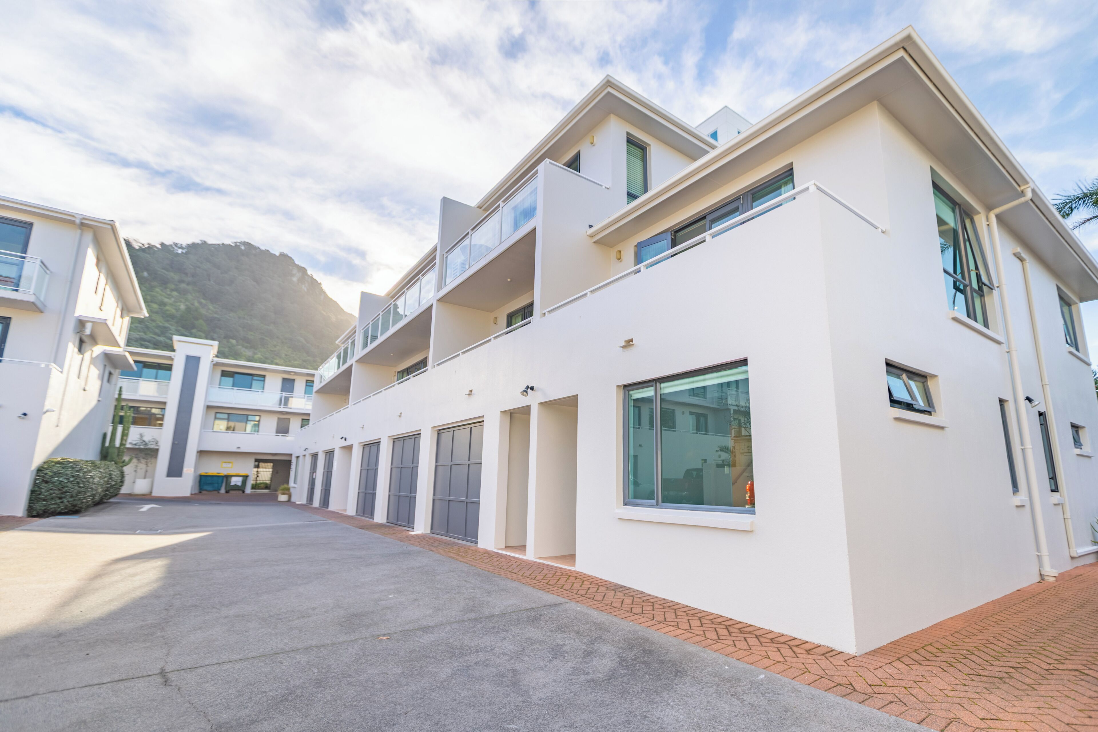 A modern multi-story residential building with a flat roof, large windows, and a paved courtyard.
