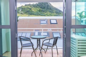 A balcony with two chairs and a table, overlooking a hillside with buildings. - Capri On Pilot Bay (Tauranga)