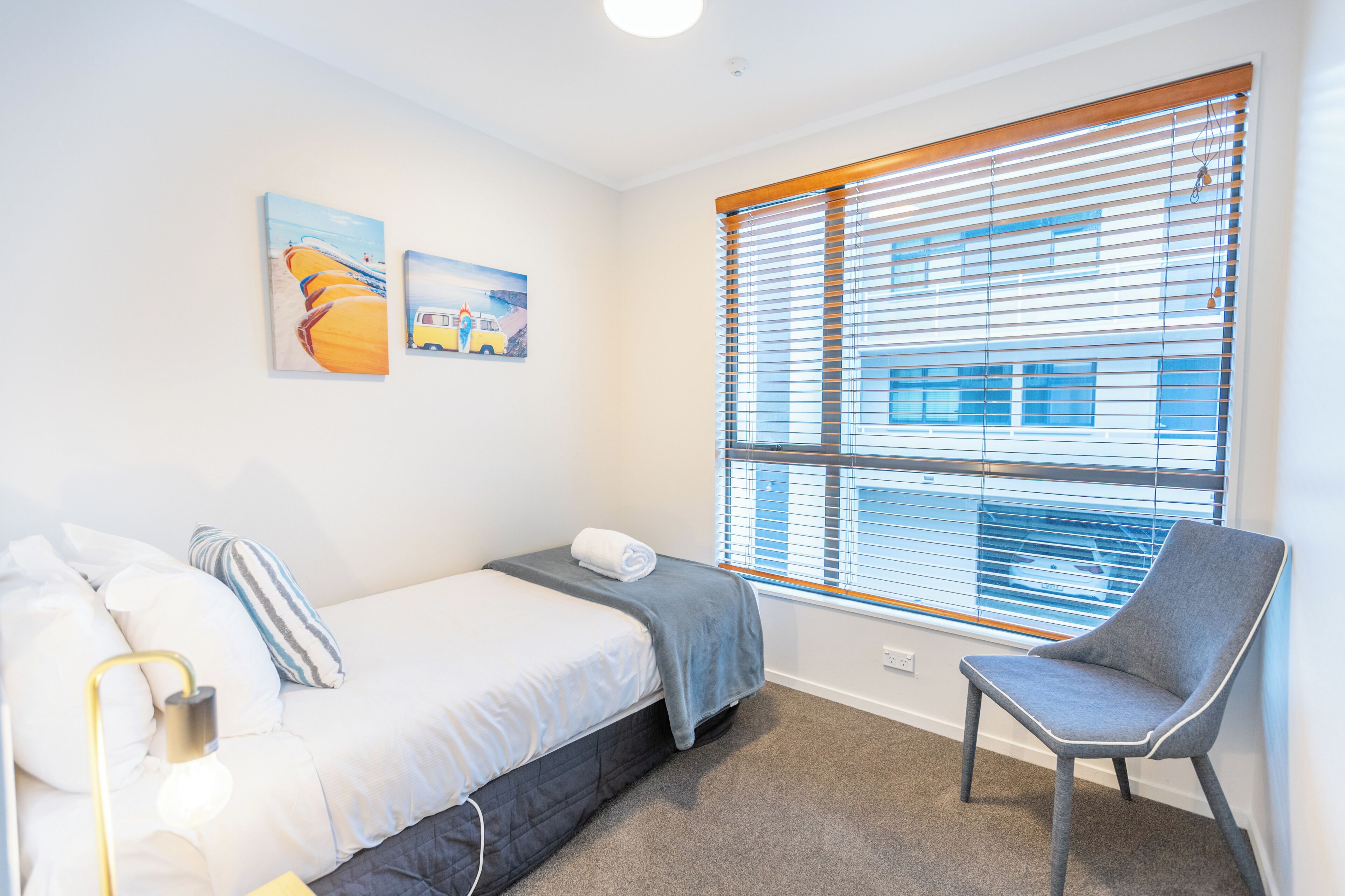 A hotel room with a bed, a chair, a window with blinds, and artwork on the wall.