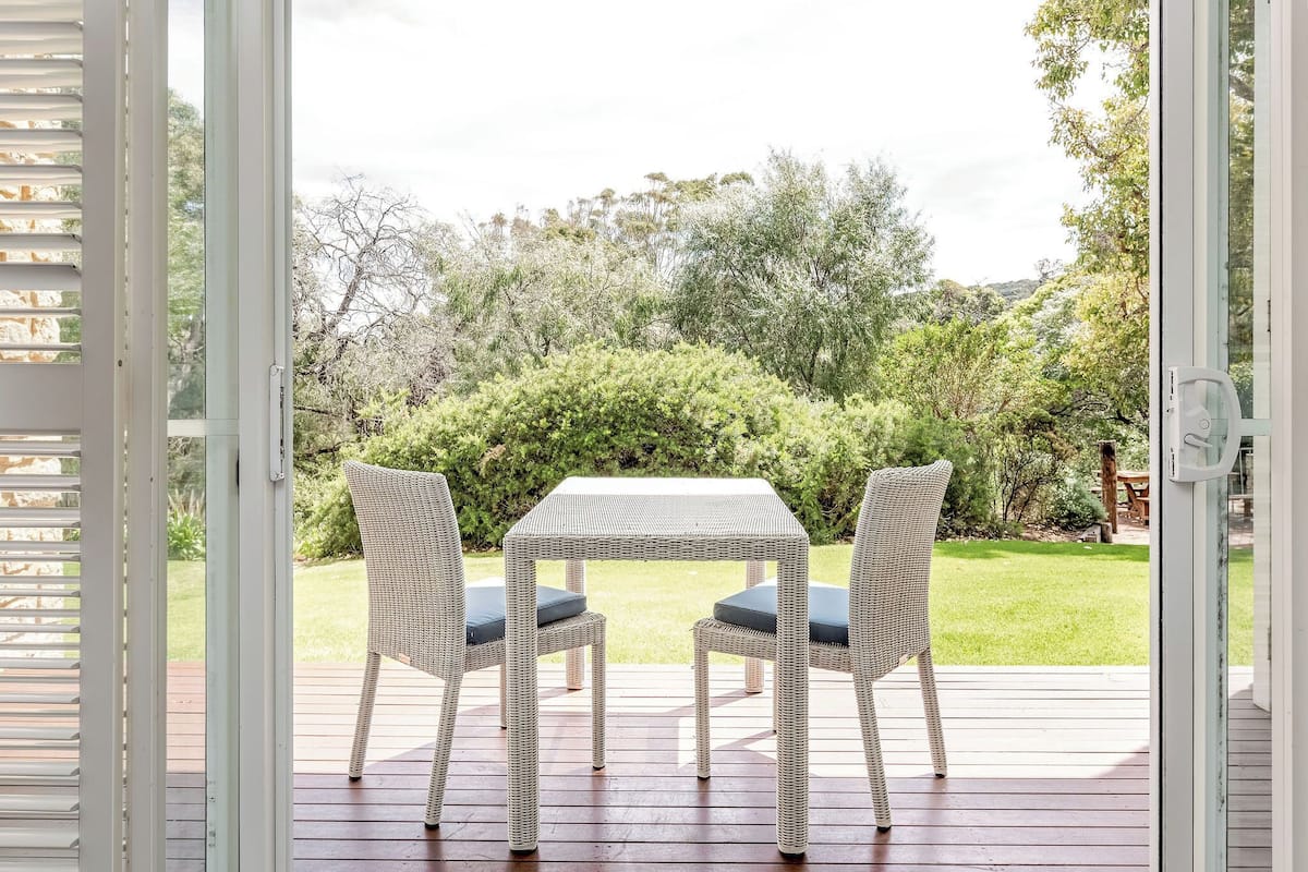 Une porte ouverte donnant sur un patio équipé d’une table et de deux chaises, le tout entouré de verdure.