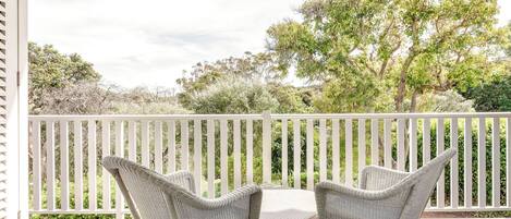 A covered outdoor seating area with wicker chairs and a table, overlooking a white fence and greenery.