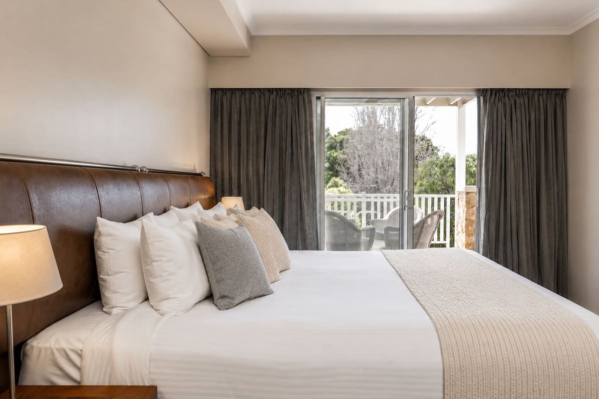 Une chambre d’hôtel avec un grand lit, une tête de lit en bois, une table de chevet avec une lampe et un balcon doté d’une balustrade blanche.