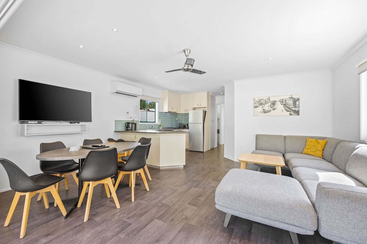 A modern living space with a dining area, kitchen, and a gray sofa with a yellow pillow.