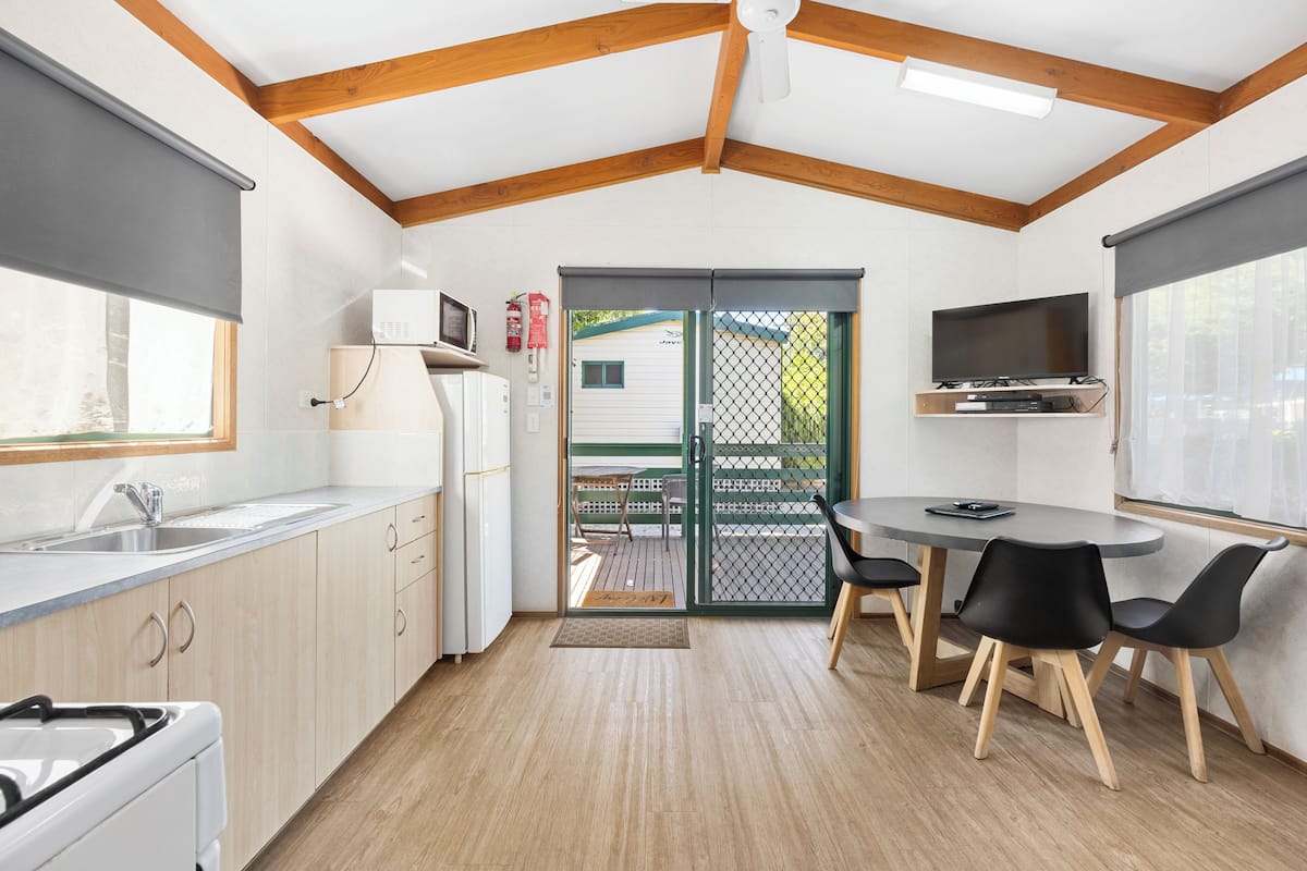 A compact kitchen with wooden beams, a dining area with a table and chairs, a TV mounted above the table, and a sliding glass door leading outside.