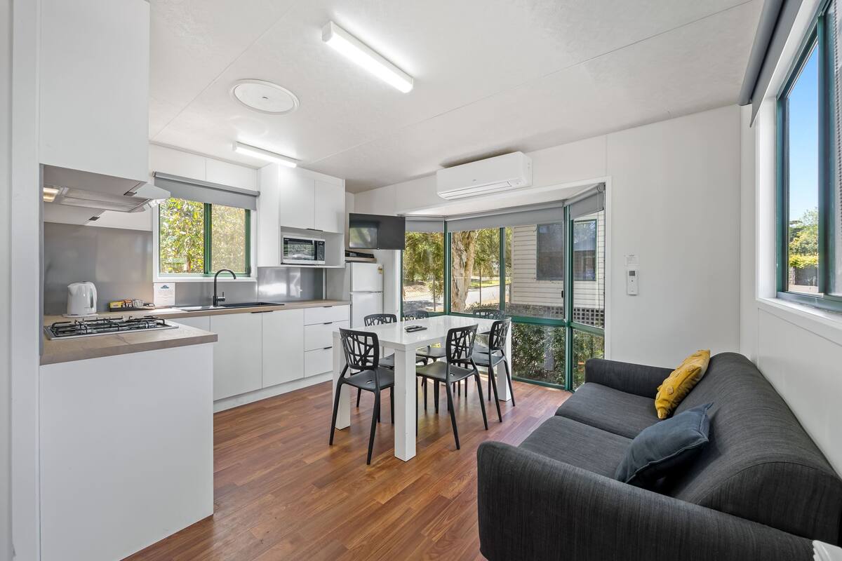 A modern kitchen with white cabinetry, a dining area with a table and chairs, and a grey sofa with a yellow pillow.