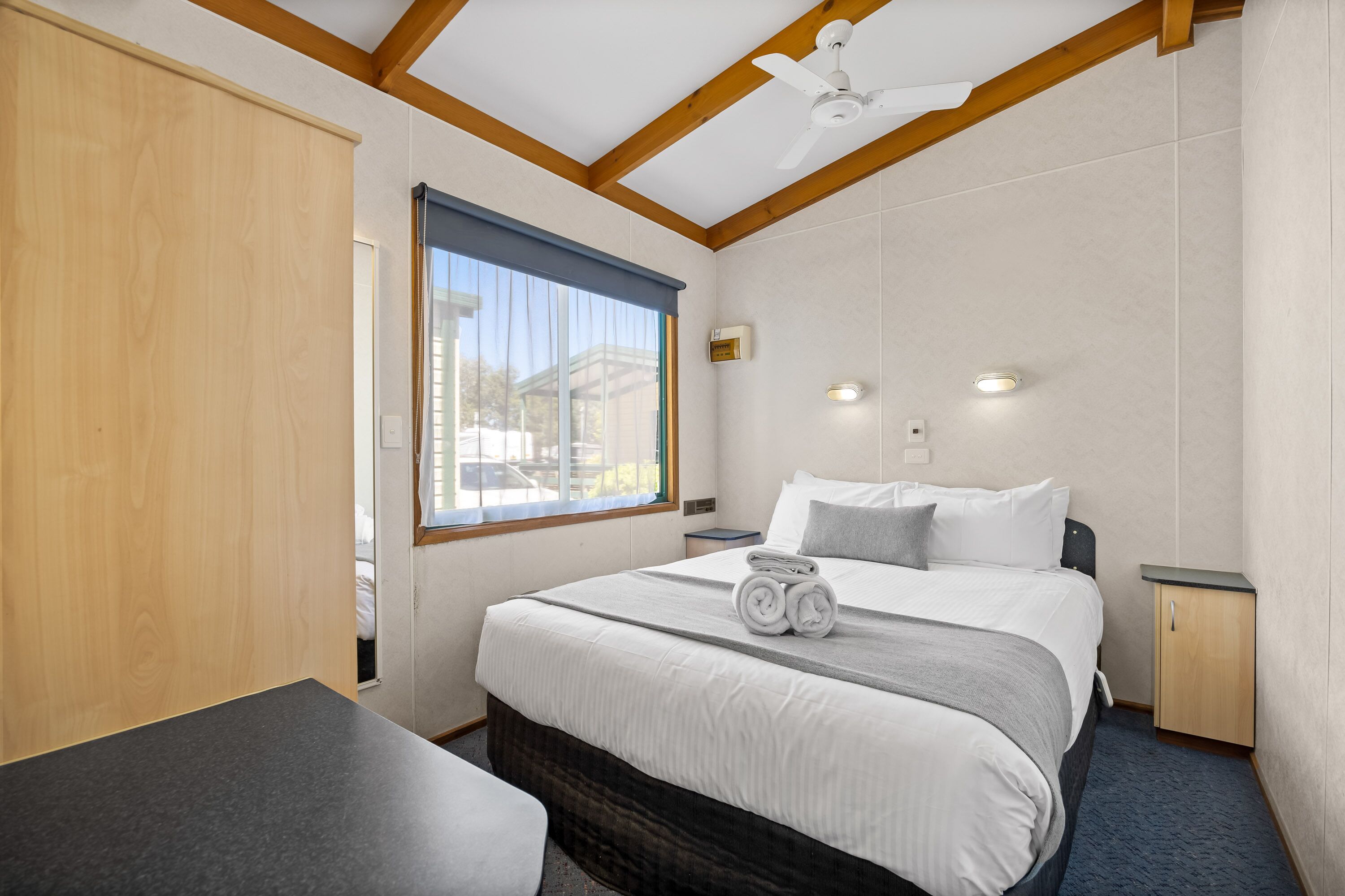 A hotel room with a bed, a bedside table, a nightstand, a window with curtains, a ceiling fan, and a view of the outside.