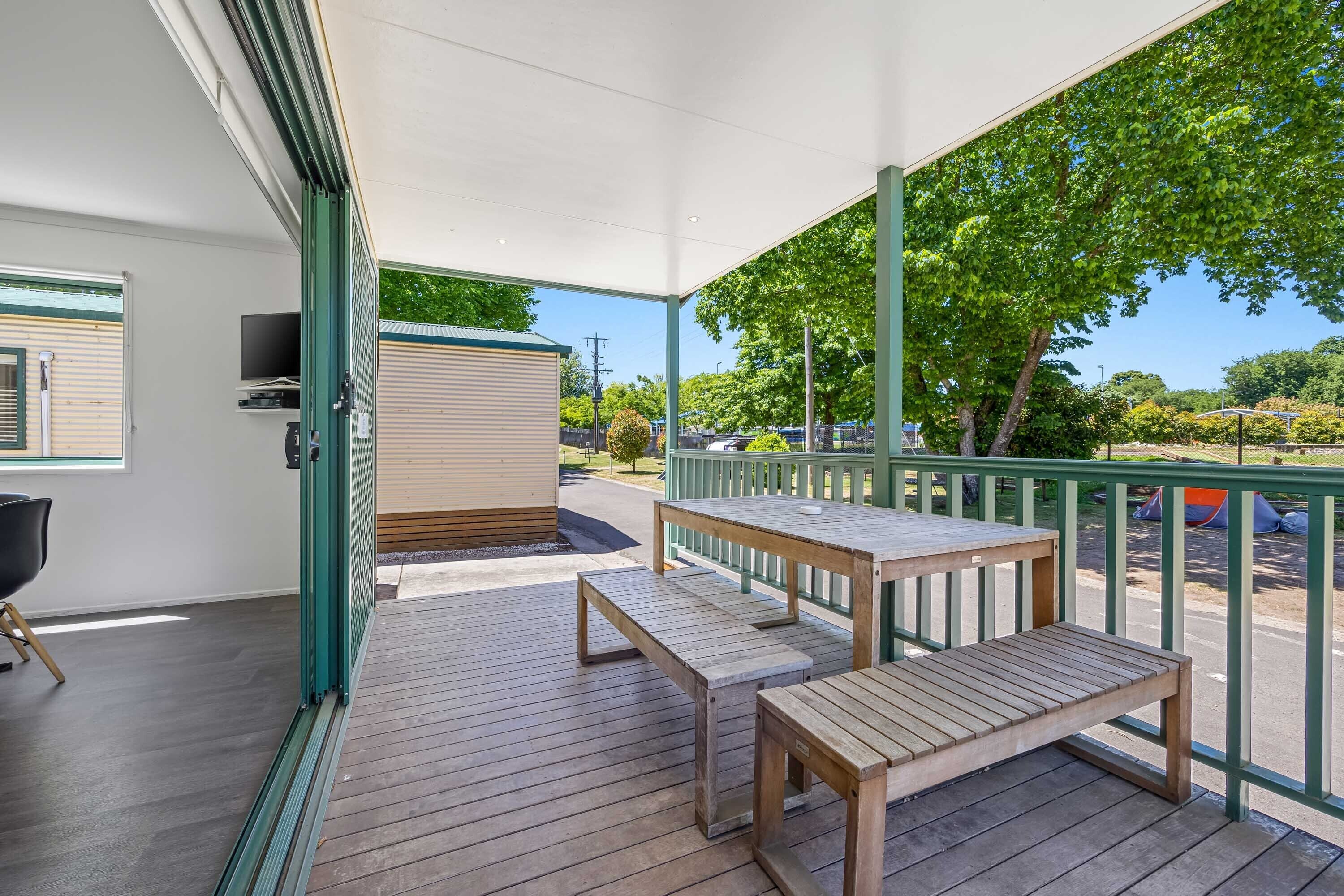 A covered outdoor area with a wooden table and benches, a TV mounted on the wall, and a view of the street and greenery.