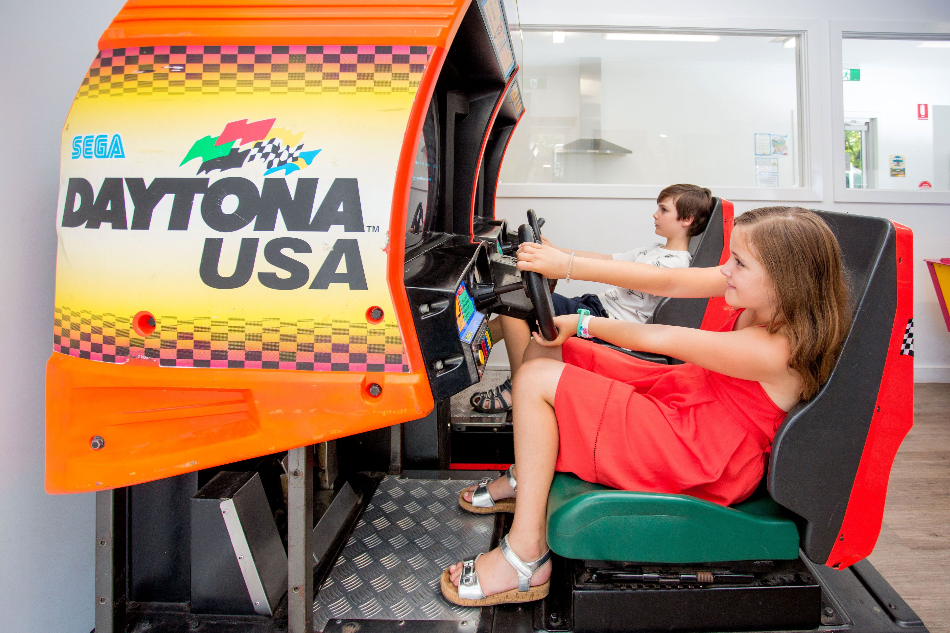 Simulatore di guida con ambientazione "Daytona USA", dotato di volante e sedile da corsa.