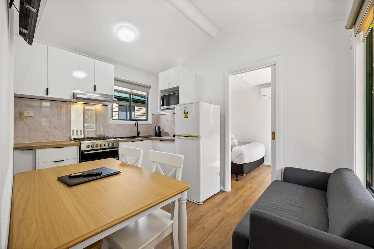 A compact kitchen with white cabinets, a stove, and a refrigerator. There is a wooden dining table with chairs and a gray sofa in the living area.
