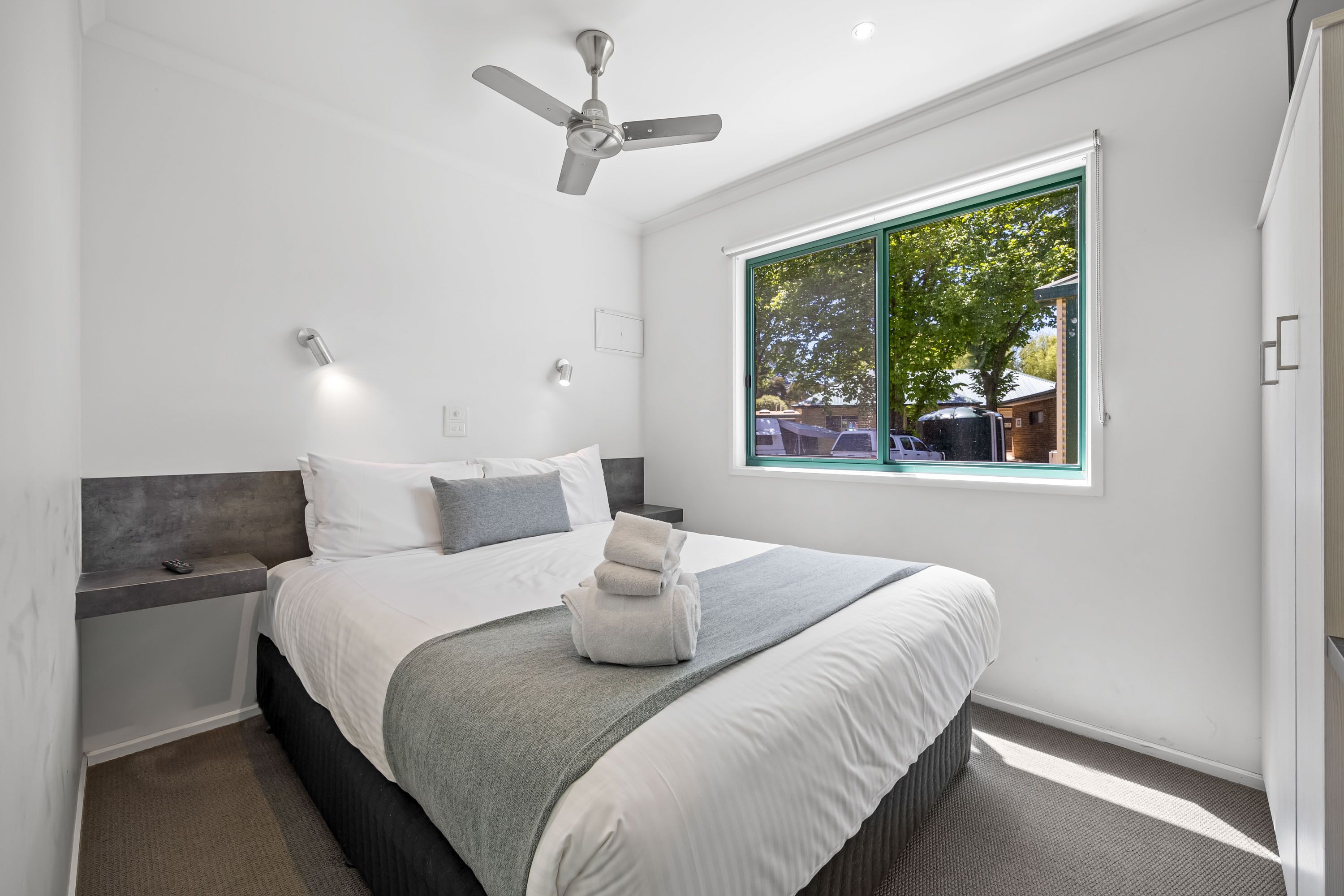 A modern bedroom with a bed, bedside tables, a ceiling fan, and a window with a view of the outside.