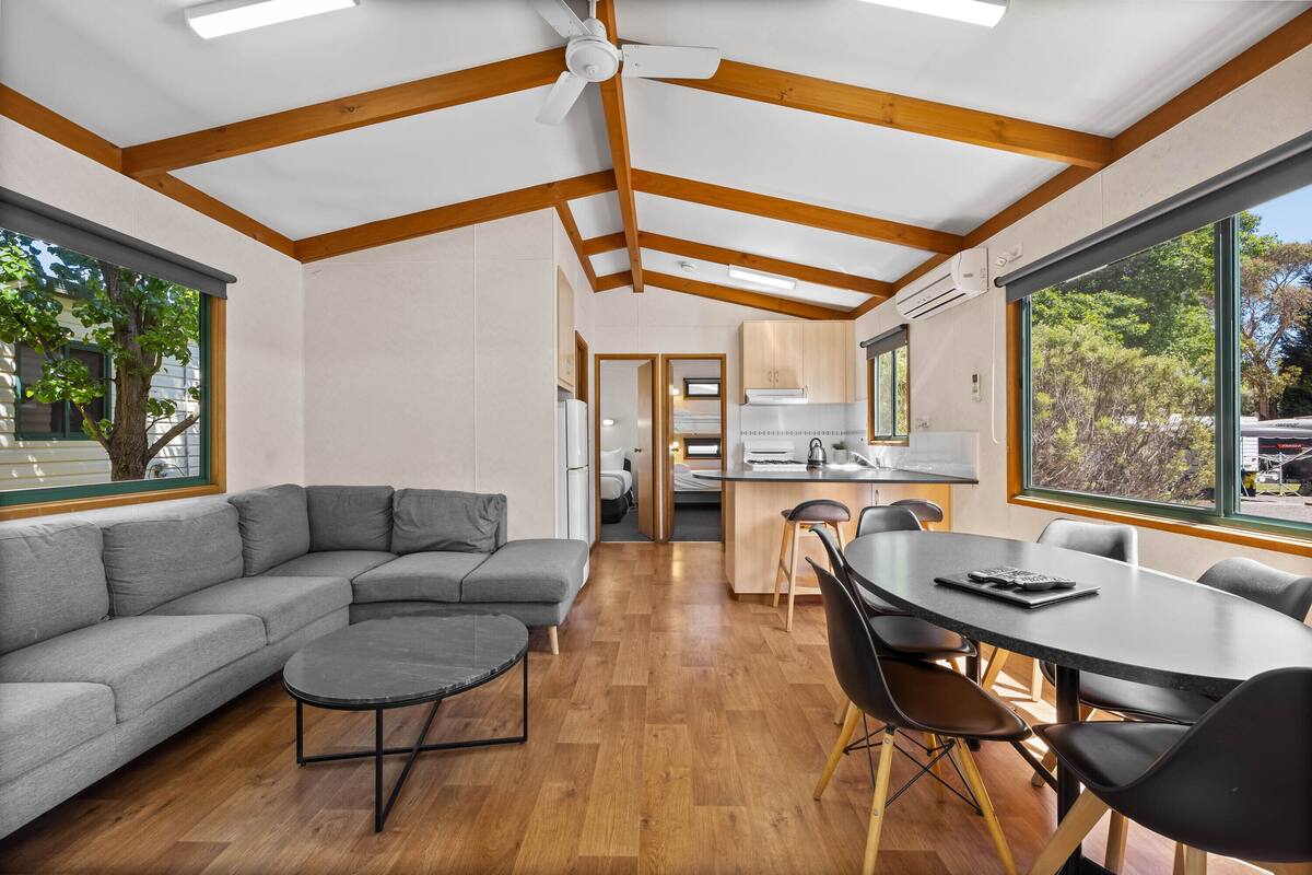 A modern interior with a sectional sofa, a round coffee table, a dining table with chairs, wooden beams on the ceiling, and large windows offering views of the outdoors.