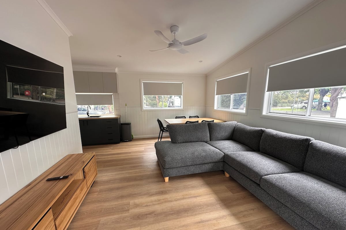 A modern living room with a grey sectional sofa, wooden flooring, a dining area with a table and chairs, and large windows with blinds.