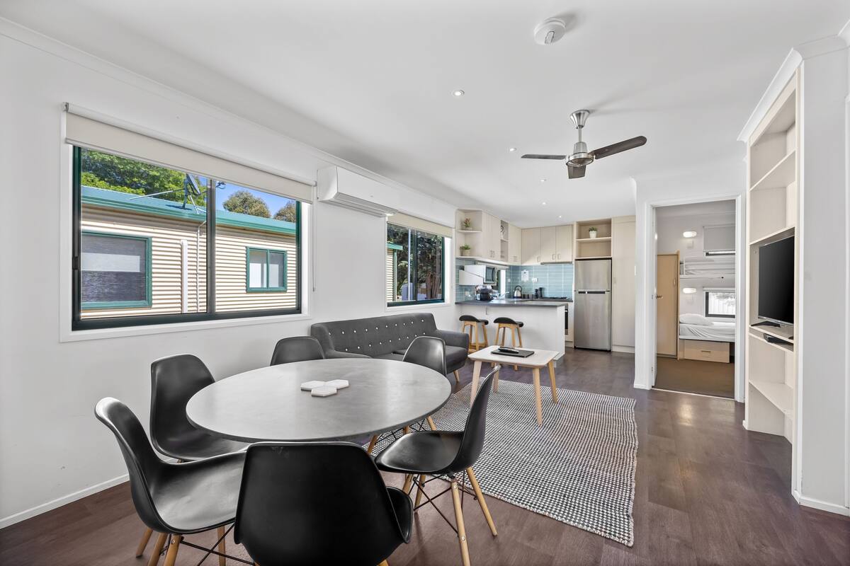 A modern kitchen with built-in appliances, a dining area with a round table and chairs, and a view of a house through large windows.