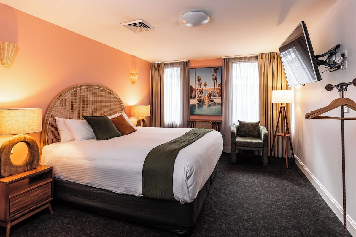 A hotel room with a large bed, a chair, a TV, and a painting on the wall.