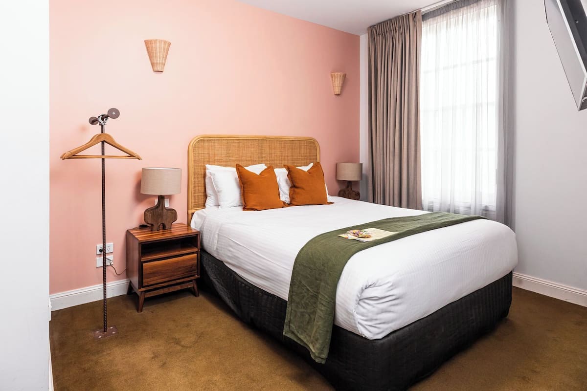 A hotel room with a large bed, a wooden nightstand, a floor lamp, and a window with curtains.