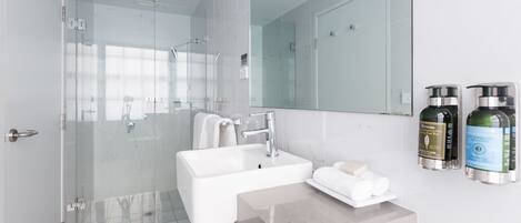 A modern bathroom with a glass-enclosed shower, a white sink, and a large mirror. There are toiletries on the counter.