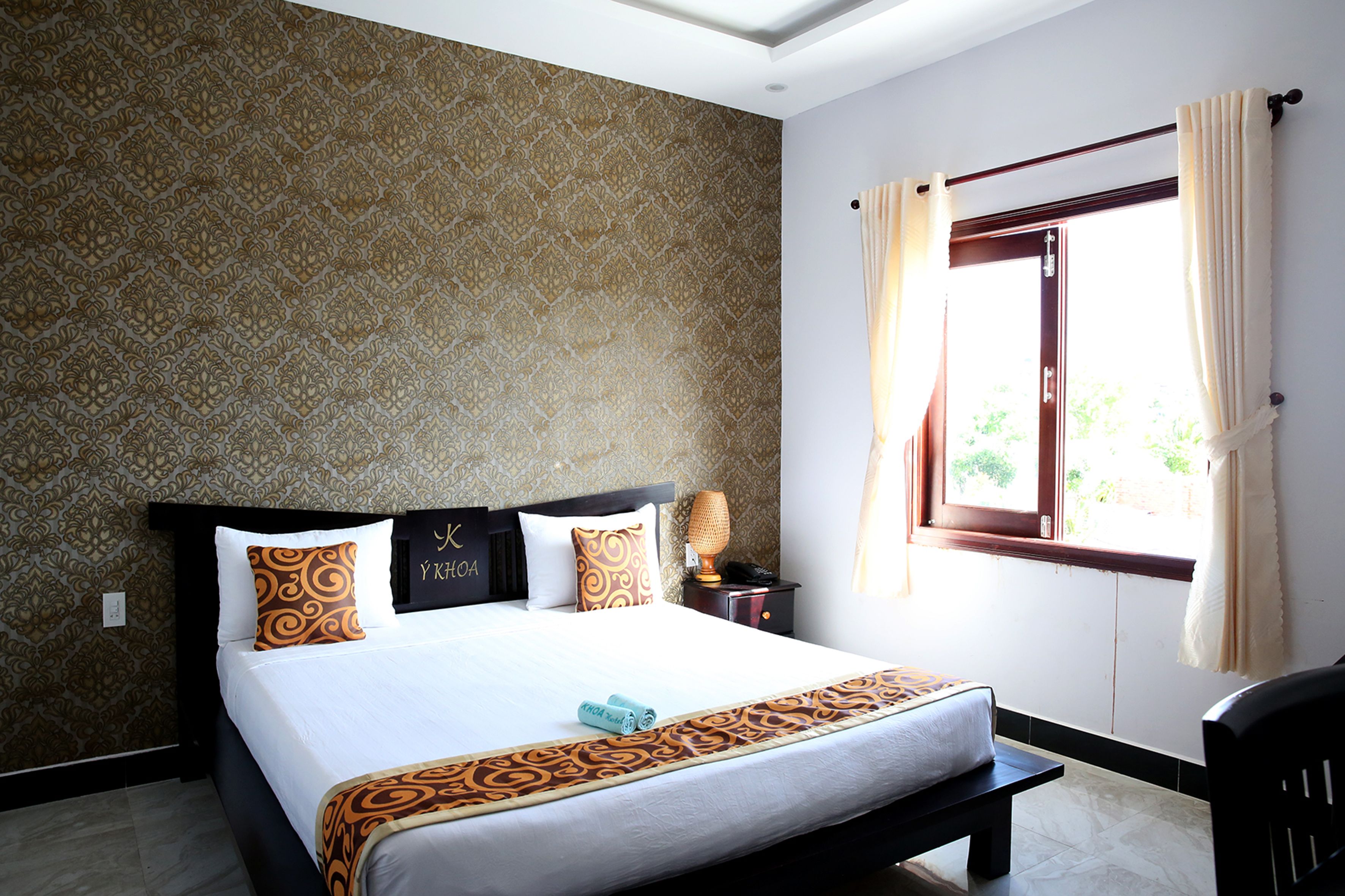A hotel room with a patterned wallpaper, a bed with a white bedspread and brown decorative pillows, a bedside table with a lamp, and a window with curtains.