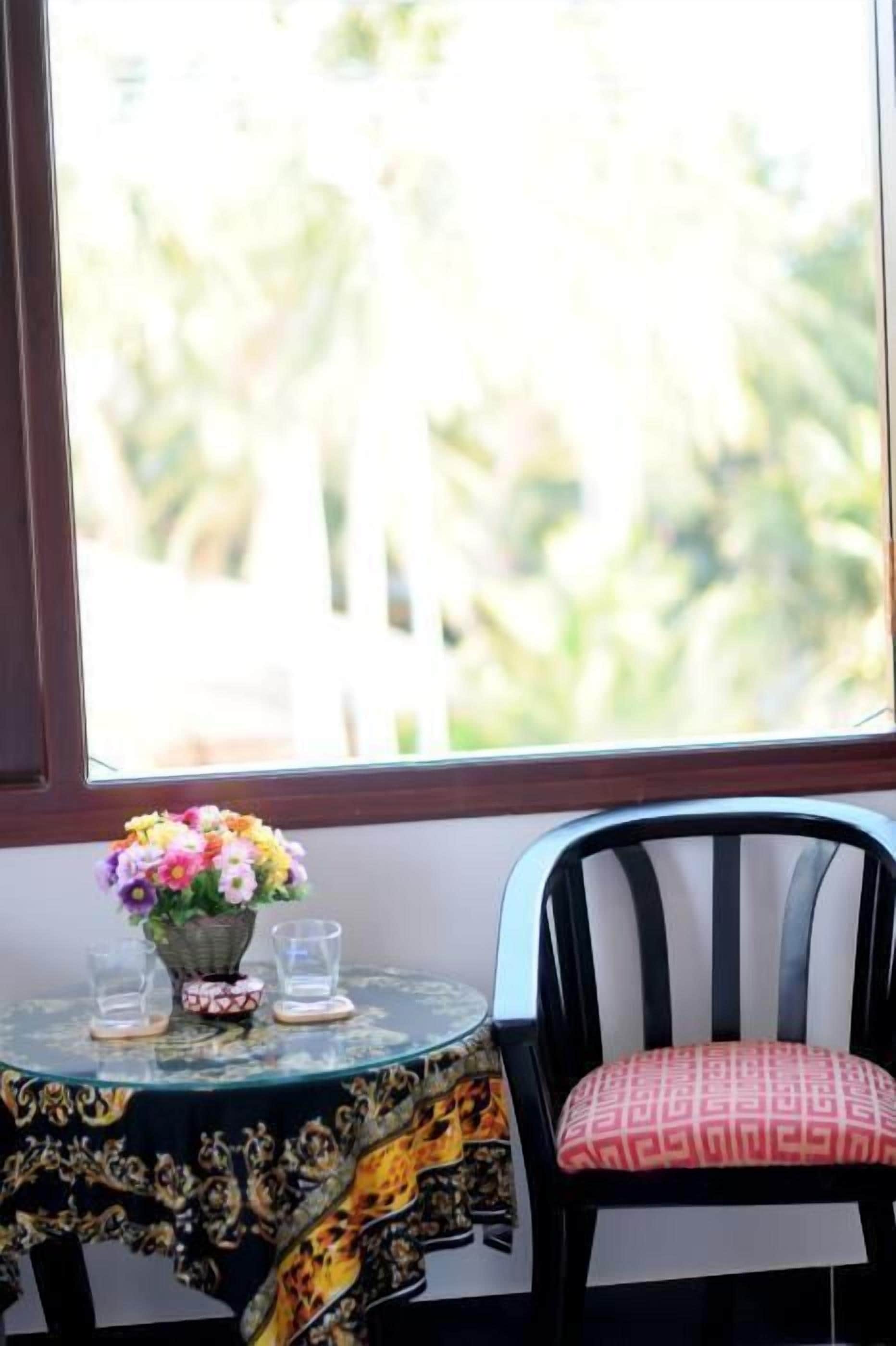 A round table with a floral centerpiece and two glasses, a chair with a patterned cushion, and a window with a view of greenery.