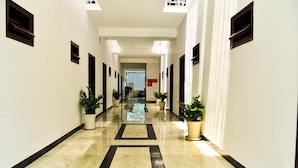 Un pasillo largo y estrecho con plantas en macetas a ambos lados, que conduce a una habitación con un ventanal grande y un letrero rojo.