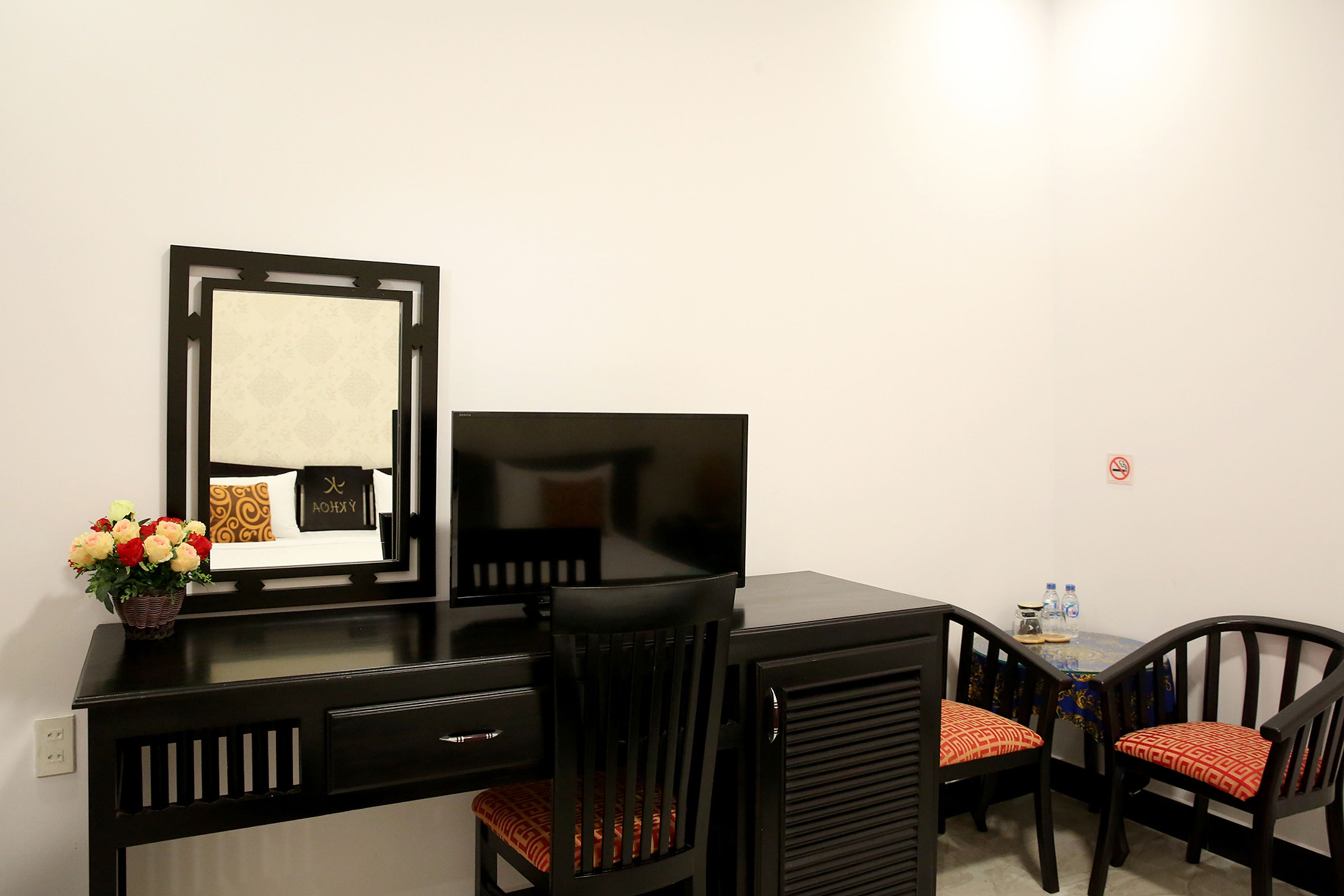 A hotel room with a black desk, a flat-screen TV, two chairs, a small table with a vase of flowers, and a wall-mounted mirror.