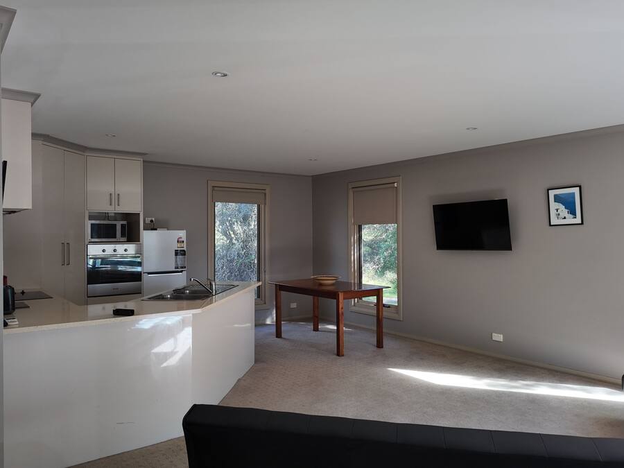 A modern kitchen with white cabinets, a microwave, and a stove. There is a dining table with a bowl on it. A black sofa is in the living area, and a TV is mounted on the wall.