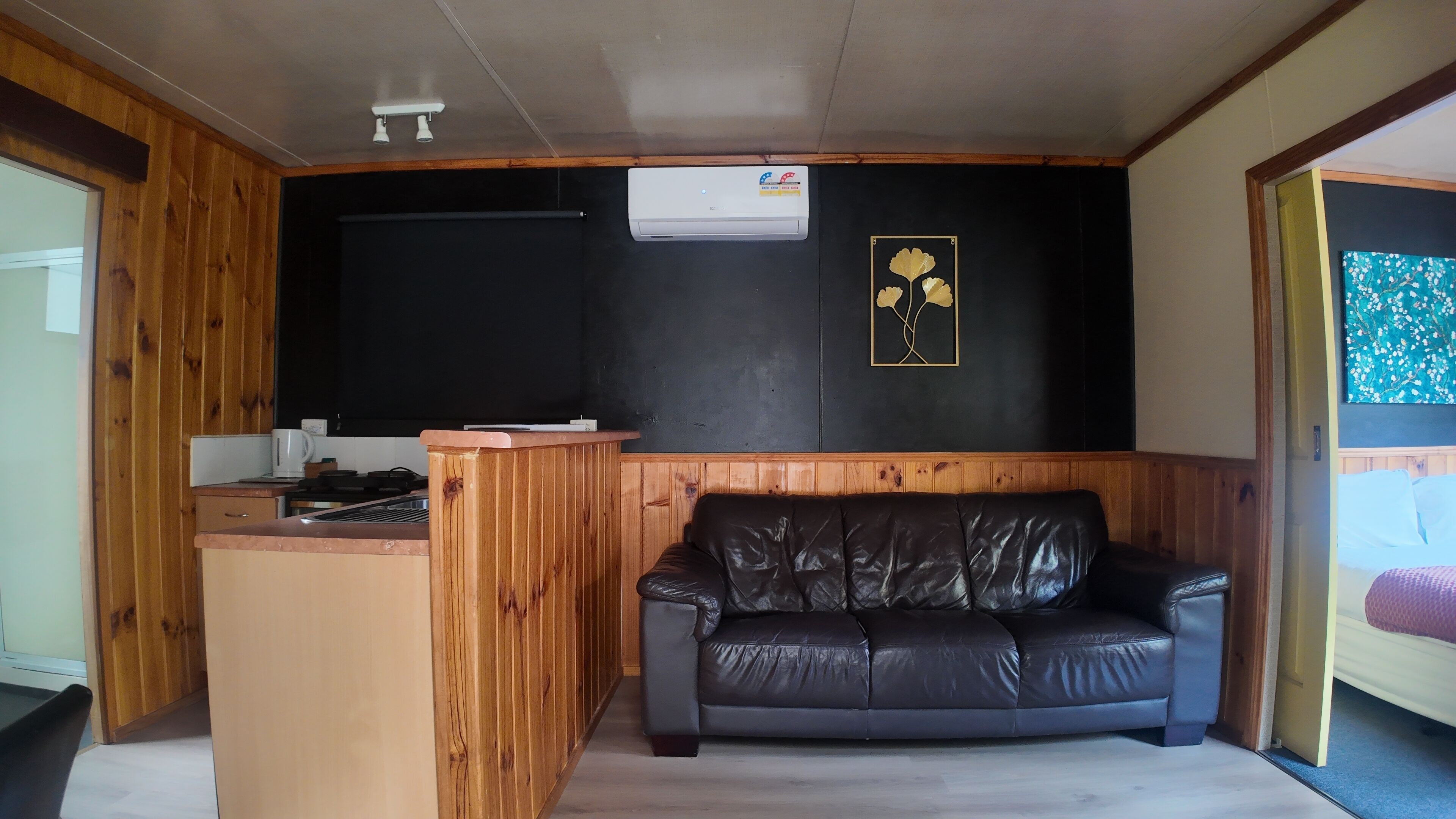 Quarto com parede de painel de madeira, sofá de couro preto, pequena cozinha com fogão e ar-condicionado instalado no teto.