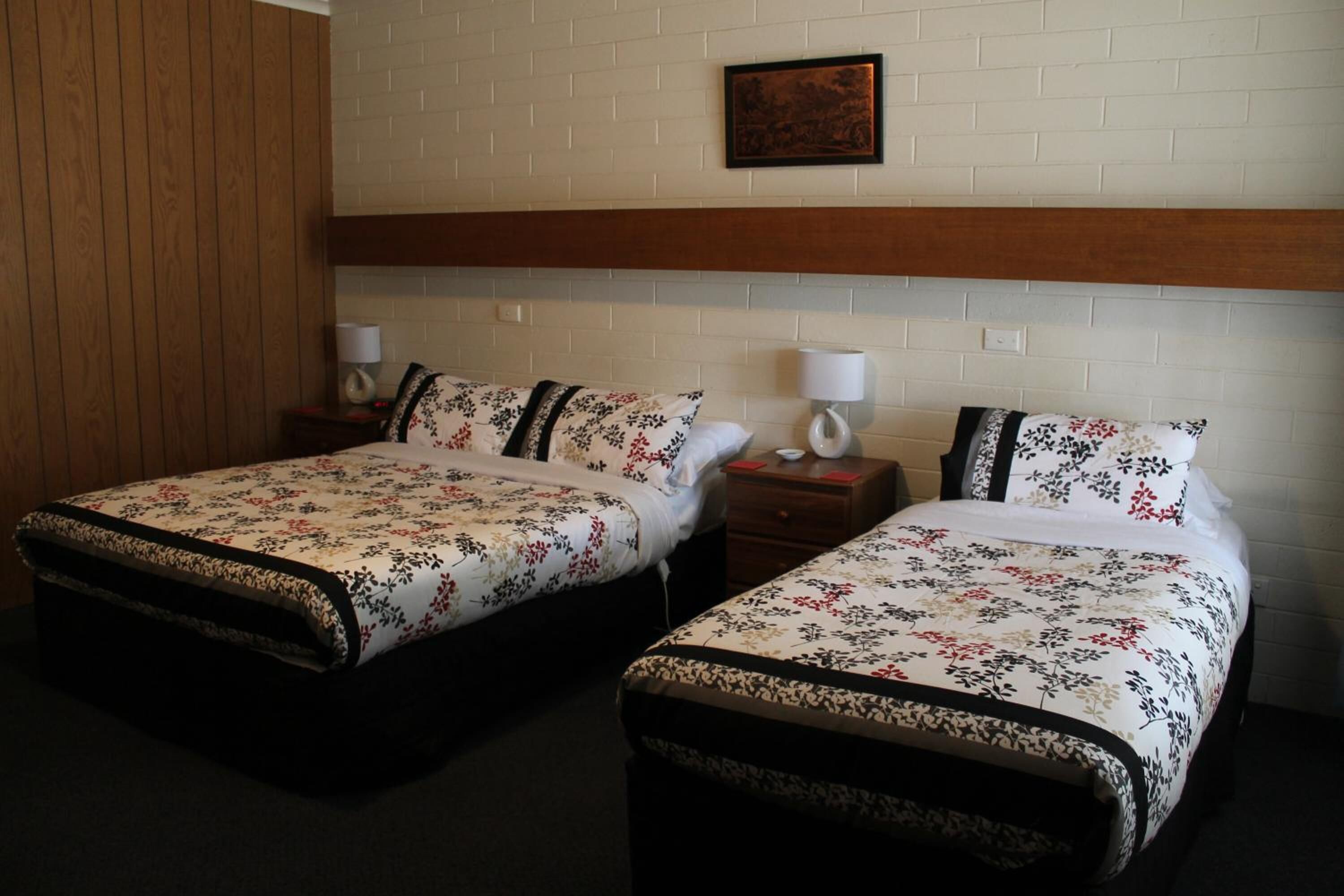 A hotel room with two single beds, each with a floral-patterned bedspread and matching pillows. There are bedside tables with lamps and a framed picture on the wall above the beds.
