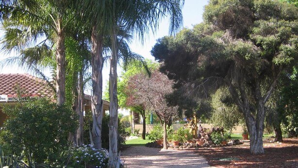 A pathway leading through a garden with various trees and shrubs, a building with a tiled roof in the background, and a sign indicating a motel. - Orana Motor Inn (Irymple)