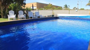 A swimming pool with clear blue water, surrounded by white lounge chairs and a wooden fence. - Orana Motor Inn (Irymple)