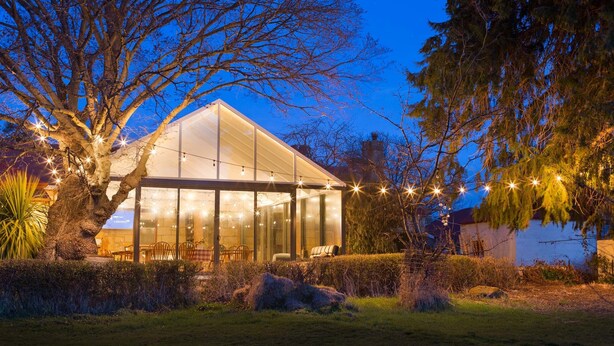 A modern glass house with a dining area and outdoor seating, surrounded by trees and a well-maintained garden. - Ratho Farm (Bothwell)