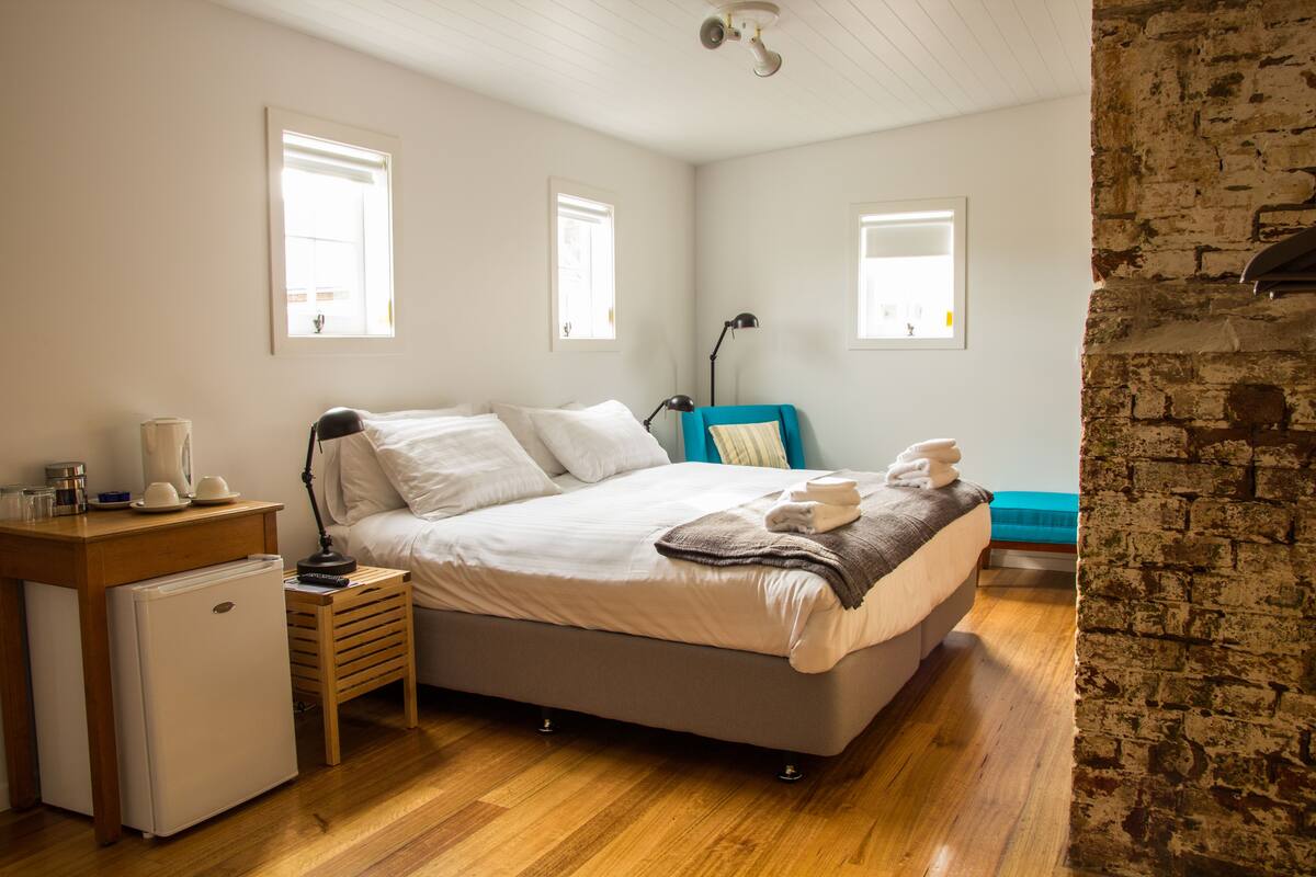A bedroom with a bed, a small refrigerator, a desk with a lamp, and a brick wall.