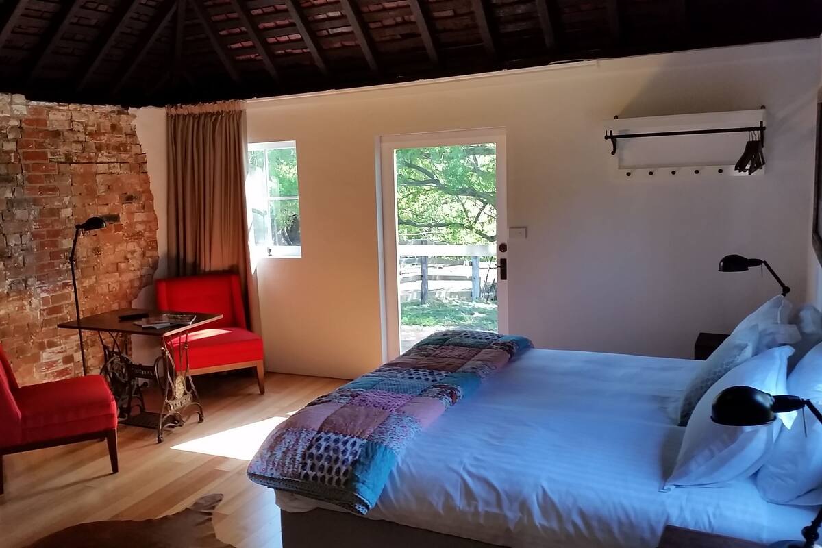A cozy room with a brick wall, a red chair, a wooden table, a bed with a quilt, and a window with a view of greenery.
