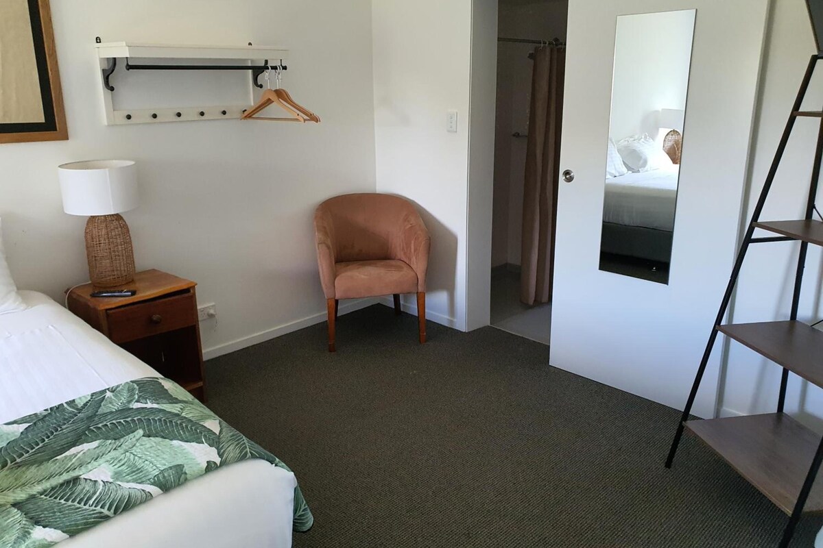 A hotel room with a bed, a chair, a nightstand with a lamp, a coat rack, and a mirror.