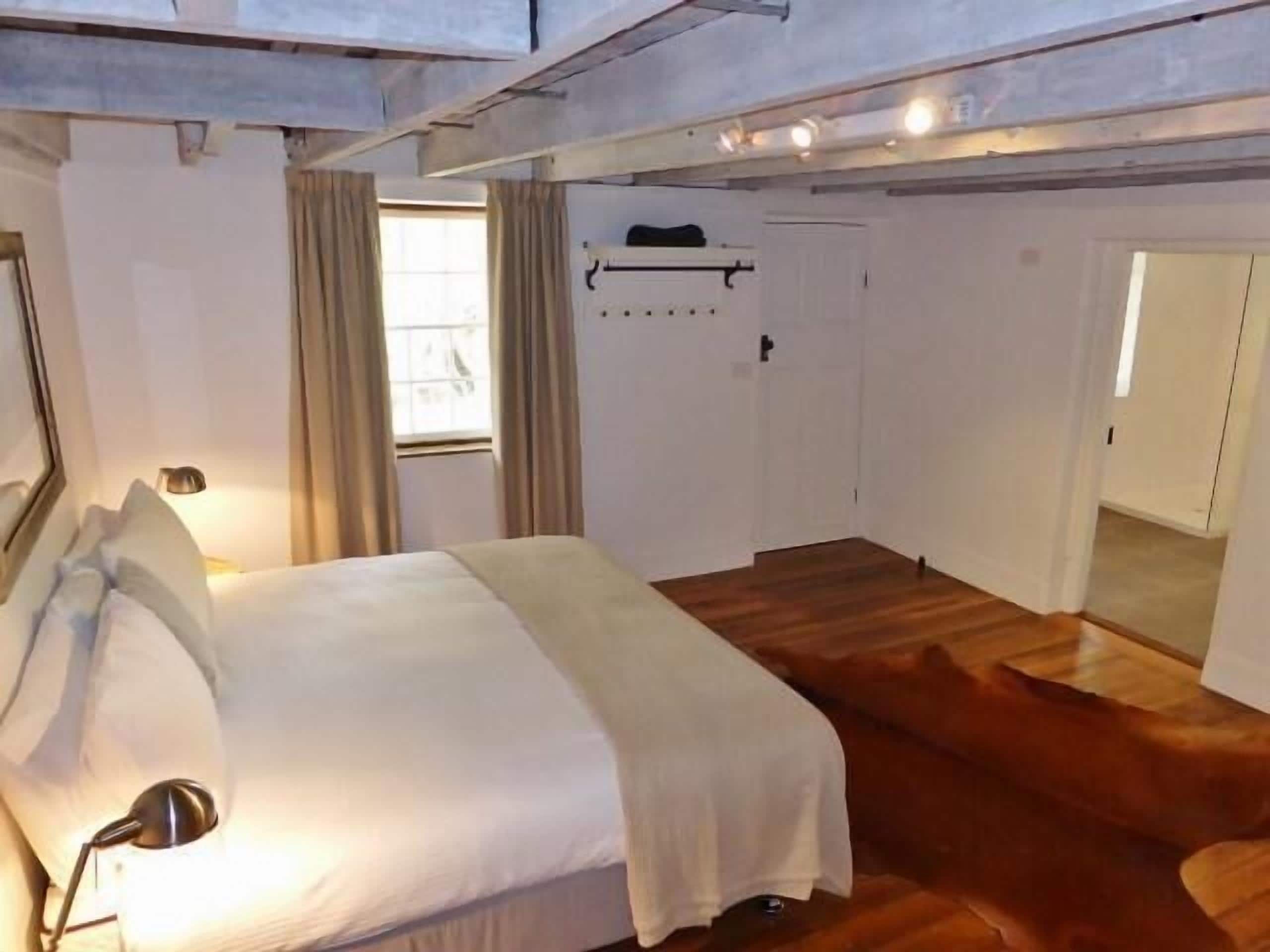 Un dormitorio con cama, lámpara de noche, ventana con cortinas, perchero y suelo de madera.