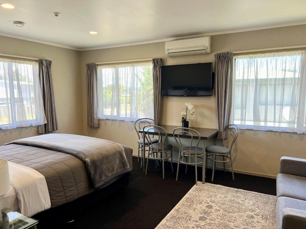 A hotel room with a bed, a desk with chairs, a television, and large windows with curtains.