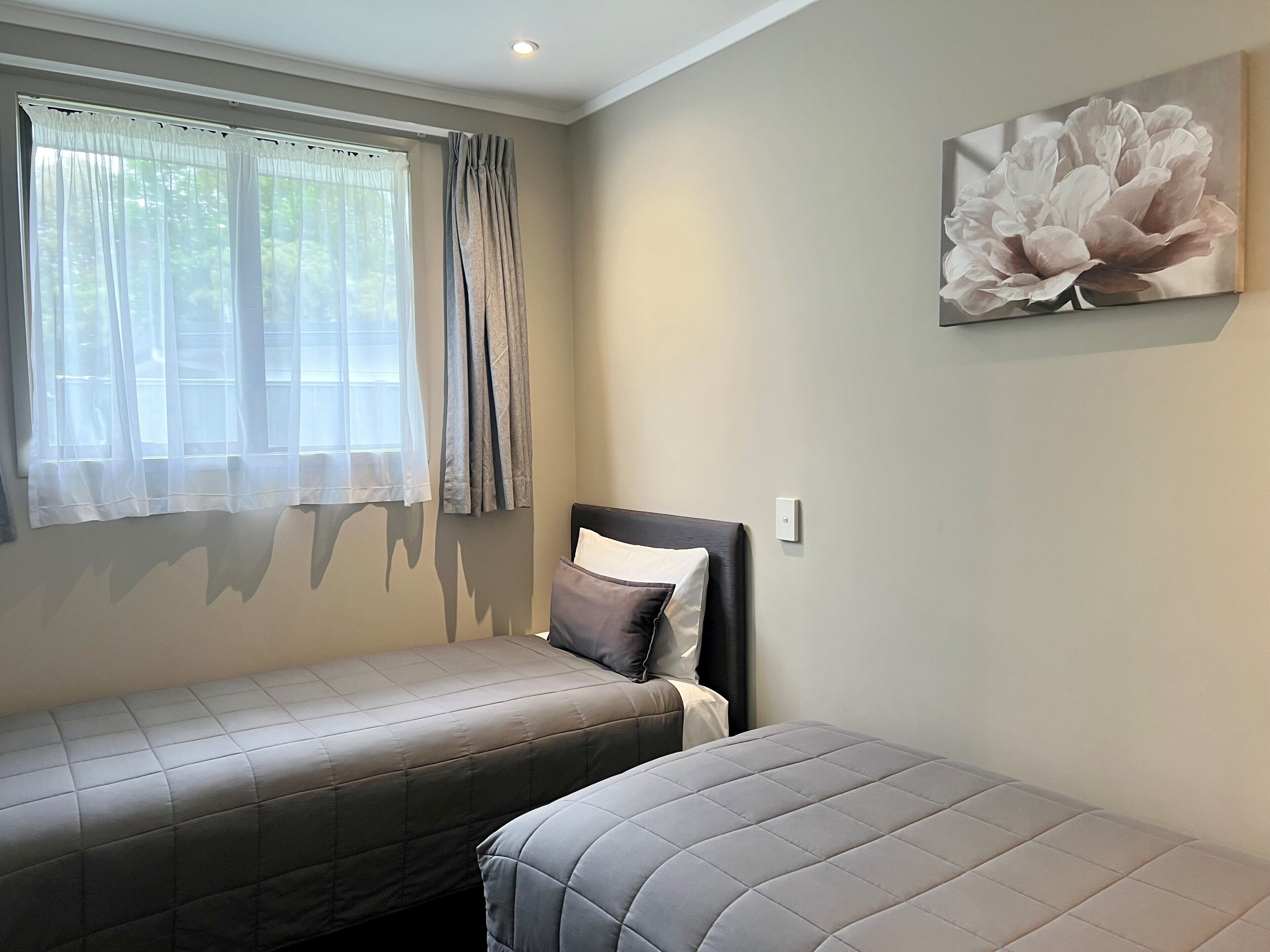A bedroom with two beds, a window with curtains, a wall-mounted light switch, and a floral painting.