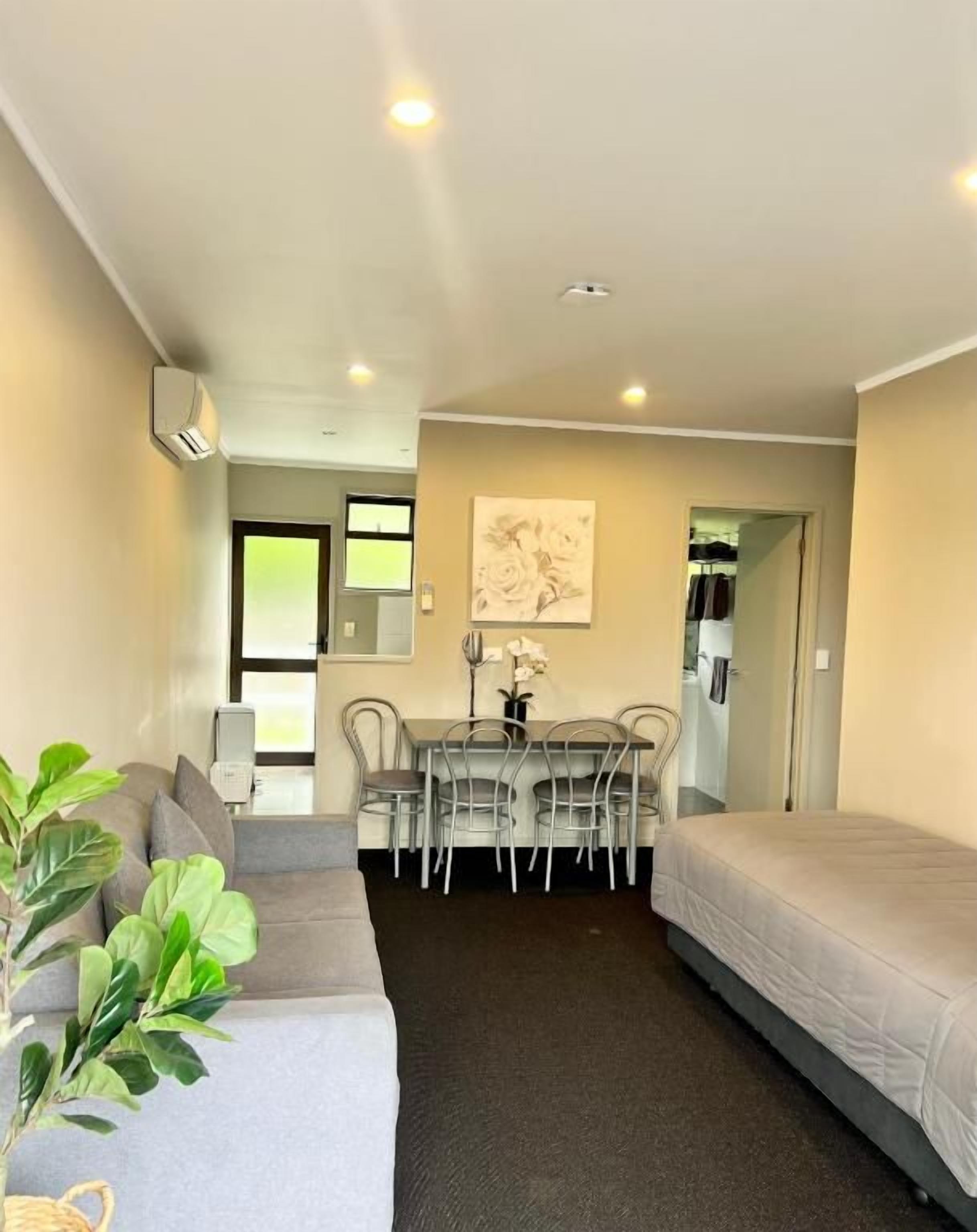 A compact hotel room with a sofa, a dining table with chairs, a bed, and a potted plant.
