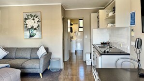 Un salon moderne avec un canapé gris, une petite table d’appoint ronde et un tableau représentant des fleurs accroché au mur. On aperçoit à l’arrière-plan un coin cuisine avec des meubles blancs, un évier et un téléphone.