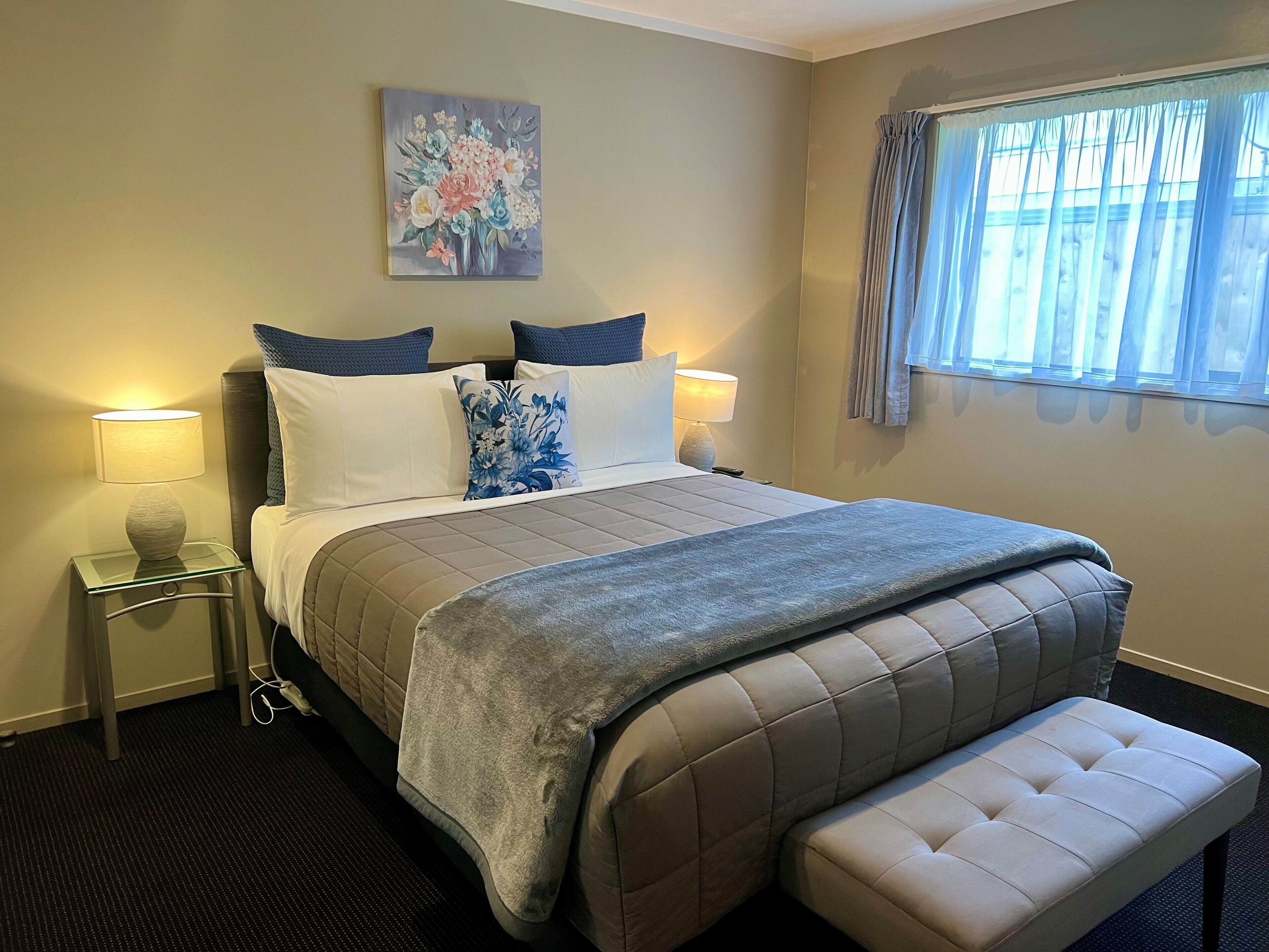 A neatly arranged bedroom with a bed, bedside lamps, a bench, and a window with curtains.